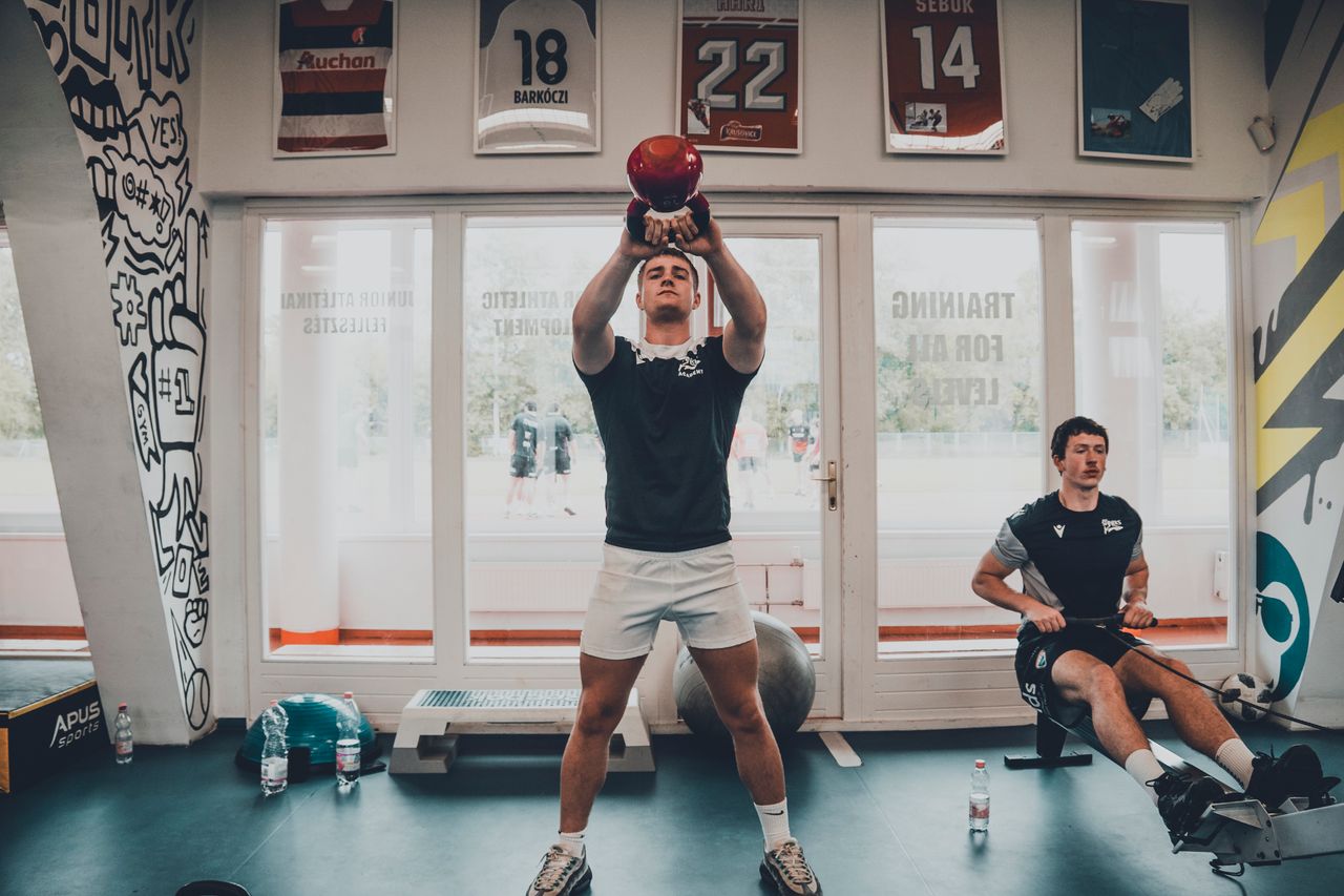 Two young men working out in a gym, focusing on strength training with kettlebells.
